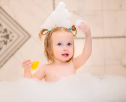 Toddler girl playing with a rubber duck in a foam-filled bathtub at home.