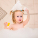 Toddler girl playing with a rubber duck in a foam-filled bathtub at home.